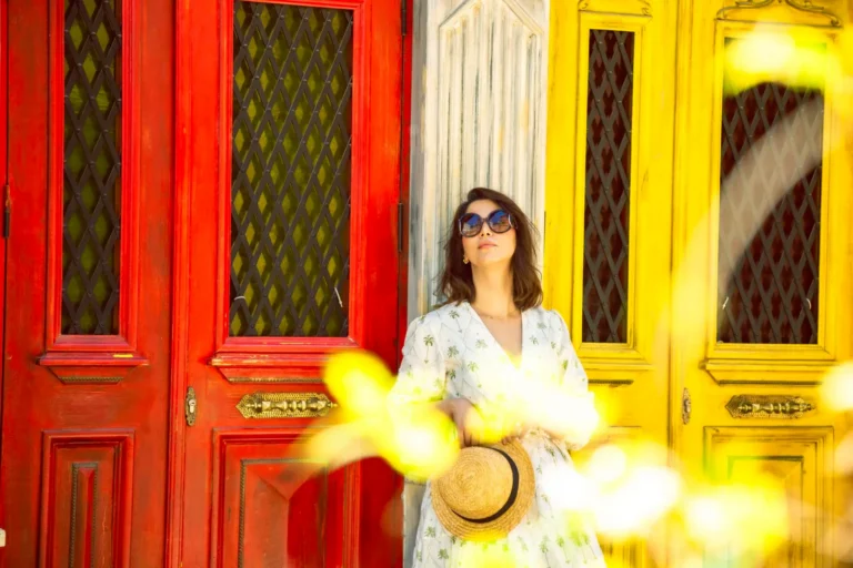 A stylish woman in sunglasses and a white dress holds a straw hat while leaning against vibrant red and yellow vintage doors.