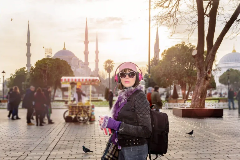 A female tourist wearing pink headphones and a purple scarf stands in a plaza in Istanbul, with the six minarets of the Blue Mosque visible in the background.