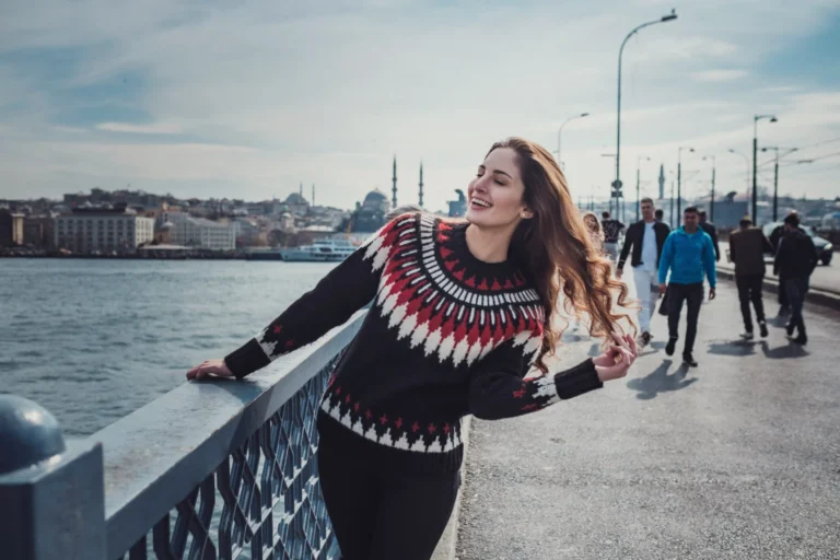 A happy young woman in a patterned sweater smiles while leaning on the railing of a bridge in Istanbul, with the city skyline visible across the water.