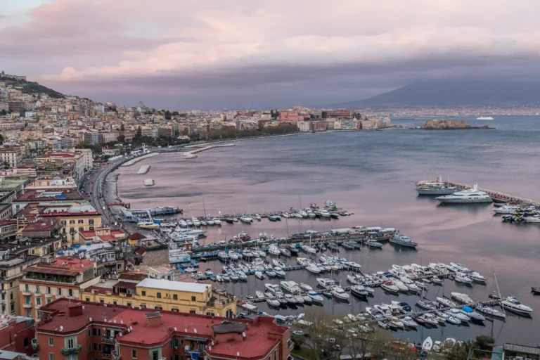 An aerial view of the Naples harbor at sunset, showing the marina full of boats, with Mount Vesuvius and the Castel dell\\\\\\\'Ovo in the distance.