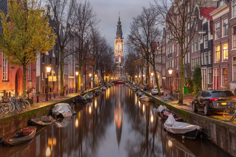 A tranquil evening view down the Groenburgwal canal in Amsterdam, with the illuminated tower of the Zuiderkerk (Southern Church) reflecting in the calm water.