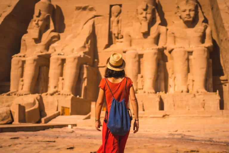 A woman in a red dress and straw hat walks towards the great rock-hewn temple of Abu Simbel, with its colossal statues of Pharaoh Ramesses II.