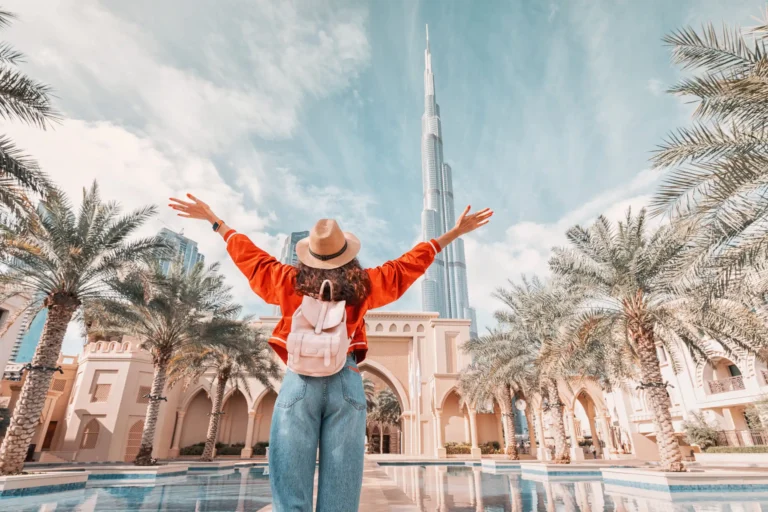 A female traveler in a hat and red jacket stands with her arms outstretched, looking towards the Burj Khalifa in Dubai from a plaza surrounded by palm trees.