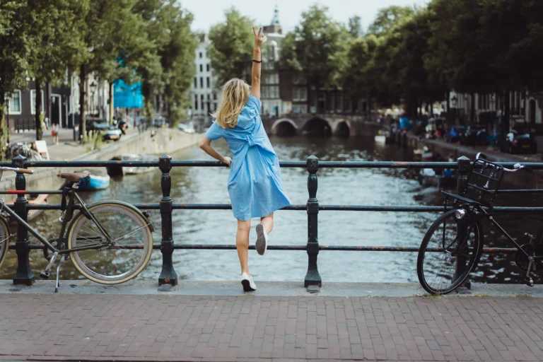 A woman in a blue dress makes a peace sign on a bridge over a canal in Amsterdam, with bicycles and historic canal houses in the background.