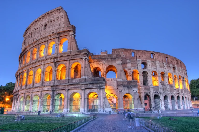 The historic Colosseum in Rome, illuminated with warm golden lights, standing against a clear, deep blue twilight sky.