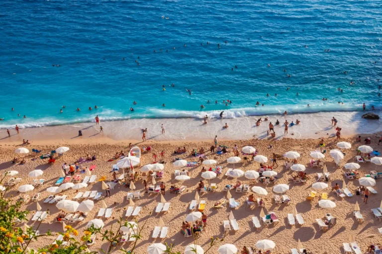 A high-angle view of the famous Kaputaş Beach in Turkey, crowded with tourists under white umbrellas on the sand and swimming in the vibrant turquoise sea.