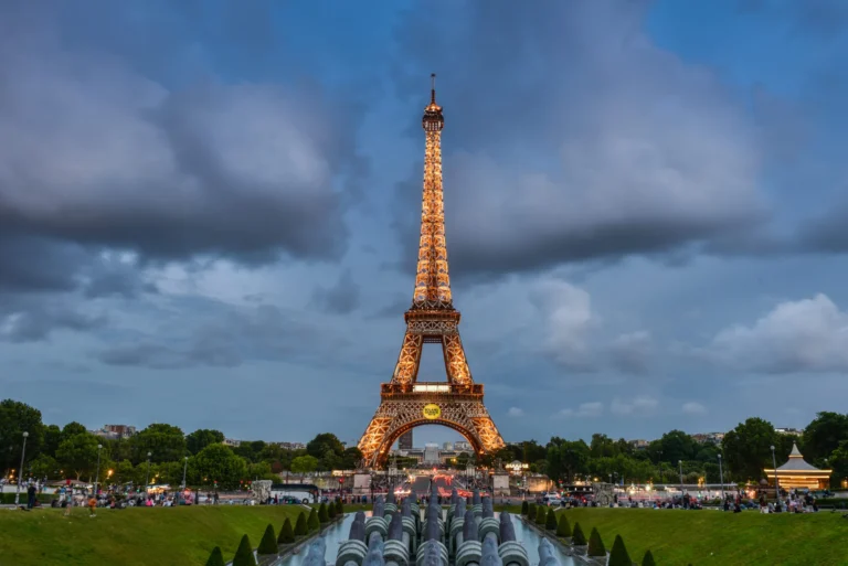 The Eiffel Tower in Paris, brilliantly illuminated with golden lights against a dramatic blue and cloudy twilight sky, as viewed from the Trocadéro Gardens.