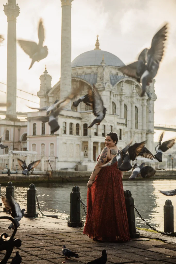 Woman in a flowing red dress at Ortaköy waterfront as pigeons fly past, with the Ortaköy Mosque glowing in soft sunrise light.