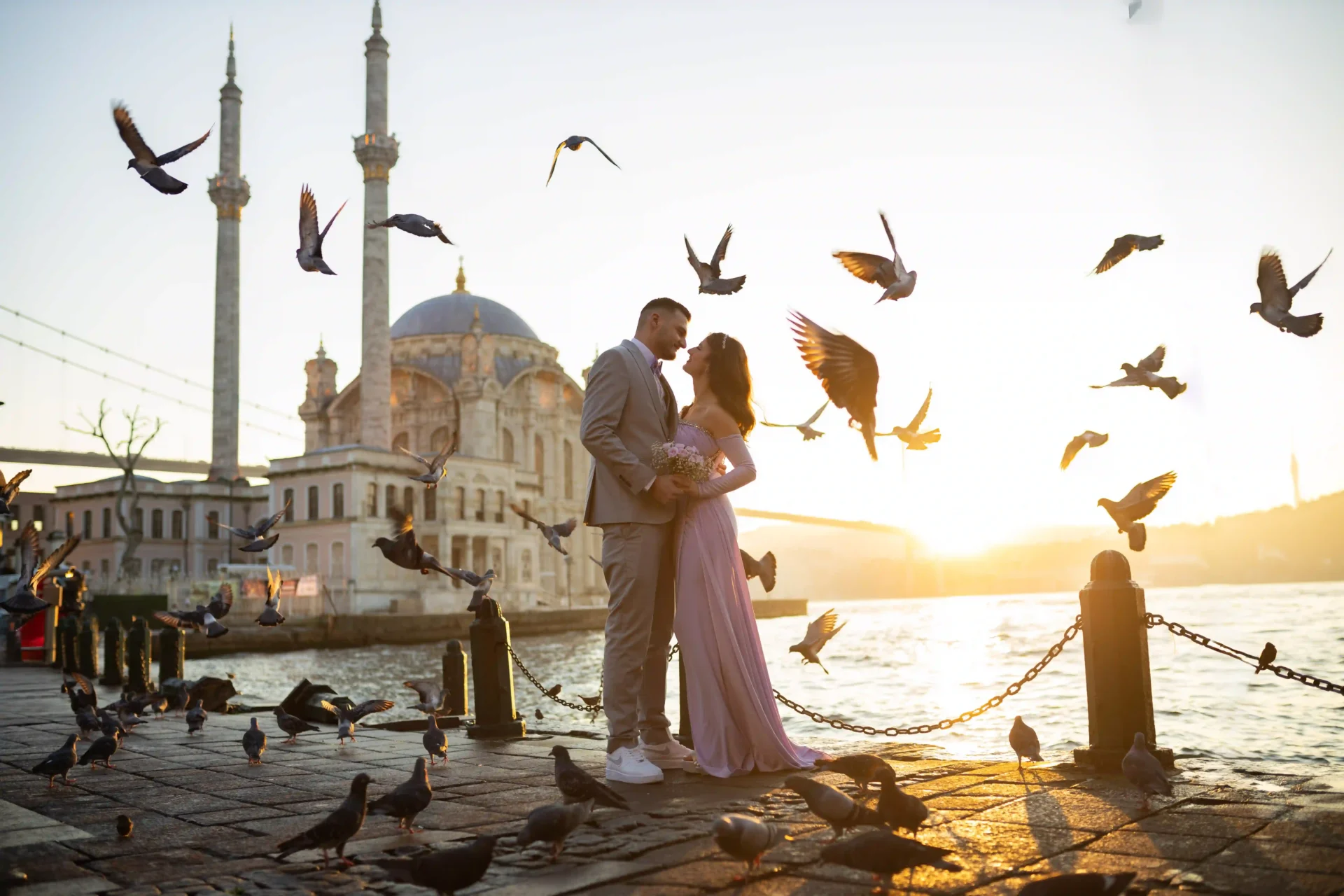 Romantic couple at Ortaköy waterfront during golden hour as pigeons fly around, with Ortaköy Mosque and the Bosphorus glowing behind.
