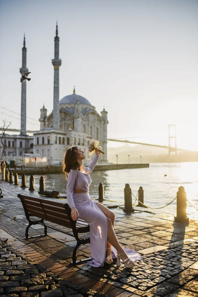 Woman in a lilac dress posing on a bench at Ortaköy waterfront at sunrise, with Ortaköy Mosque and the Bosphorus Bridge in the background.