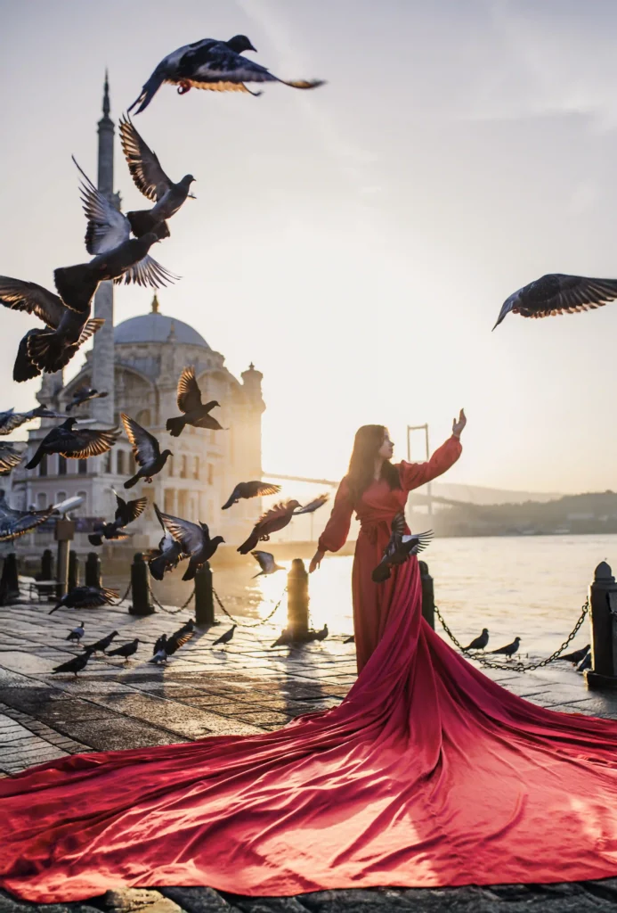 Woman in a dramatic red flying dress at Ortaköy waterfront at sunrise as pigeons fly overhead, with Ortaköy Mosque and the Bosphorus Bridge behind.