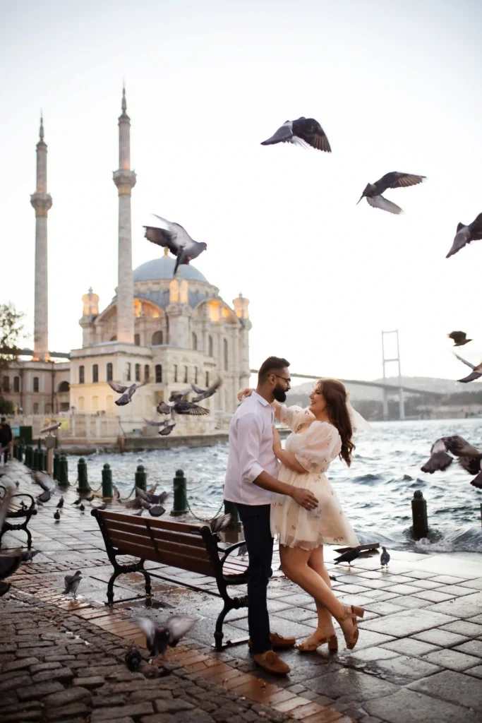 Couple embracing on Ortaköy’s Bosphorus promenade as pigeons fly past, with Ortaköy Mosque and the Bosphorus Bridge in the background.