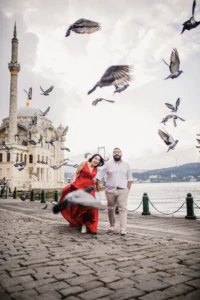 Joyful couple in Ortaköy walking along the cobblestone waterfront as pigeons fly around them, with Ortaköy Mosque and the Bosphorus in the background.