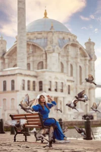 Woman in a blue dress sitting on a bench with pigeons flying around in front of Ortaköy Mosque on the Bosphorus in Istanbul, Turkey.