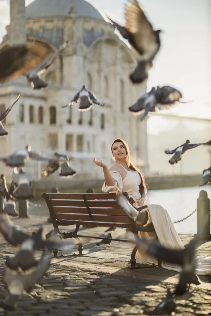 Woman on a bench near Ortaköy Mosque with pigeons flying in warm sunset light.