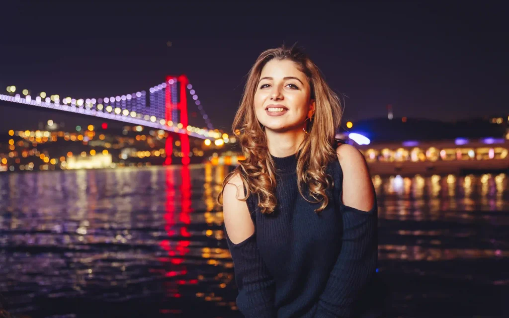 Smiling woman posing by the Bosphorus at night with the illuminated Bosphorus Bridge and city lights reflecting on the water.