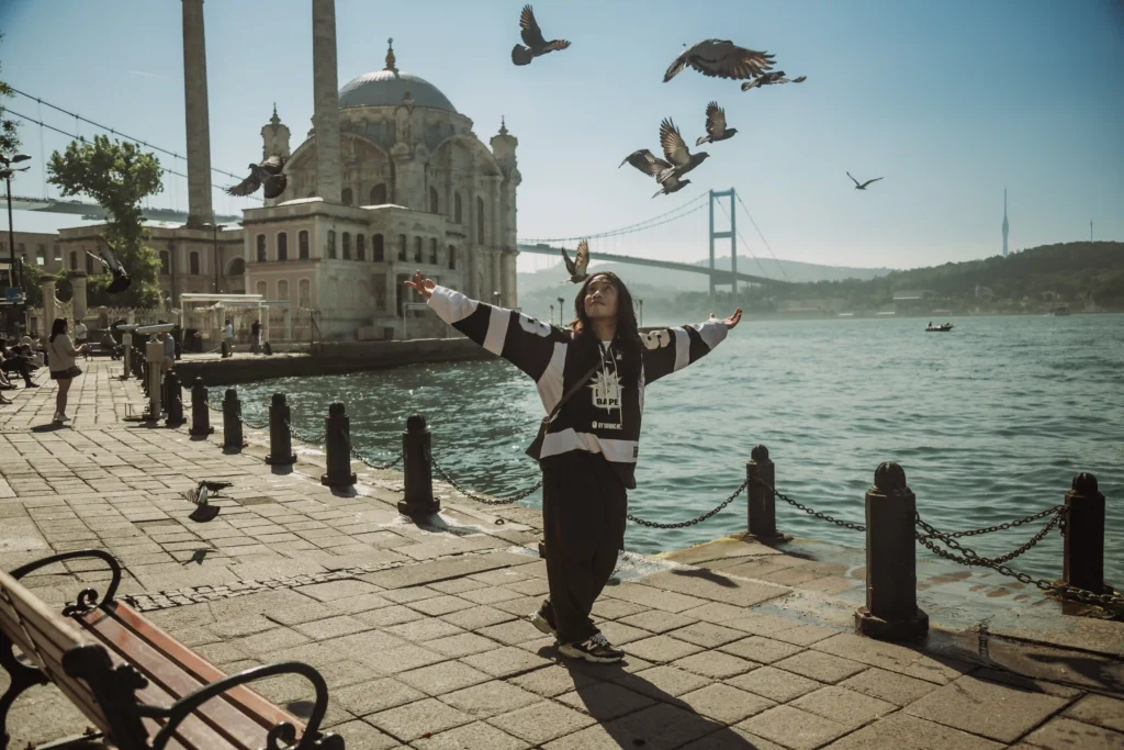 Asian man in streetwear posing with open arms near Ortaköy Mosque and Bosphorus Bridge, birds flying at sunrise in Istanbul